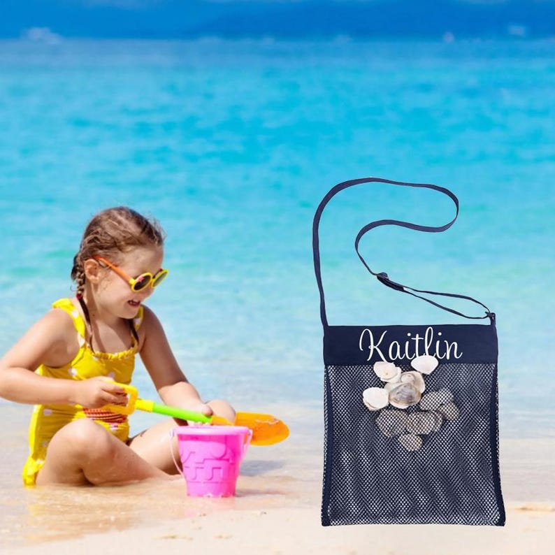 May include: Navy blue mesh beach bag with the name "Kaitlin" in white script. The bag is filled with seashells. A child in a yellow swimsuit and sunglasses plays with a pink bucket and sand toys on the beach.