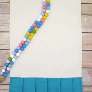 May include: A beige linen table runner with a teal blue ruffled trim. A colorful beaded garland with pastel and yellow beads is draped over the top of the runner.