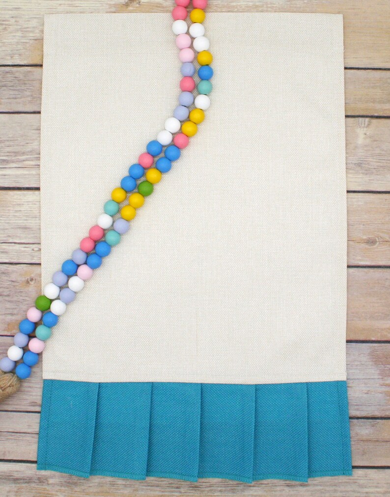 May include: A beige linen table runner with a teal blue ruffled trim. A colorful beaded garland with pastel and yellow beads is draped over the top of the runner.