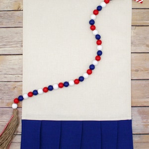 May include: A white linen kitchen towel with a blue ruffled bottom and a red, white, and blue beaded garland with a small American flag heart at the end.