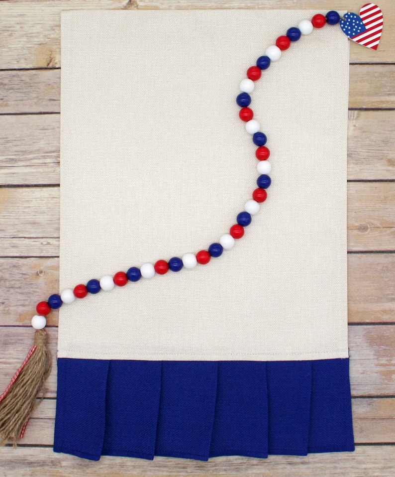 May include: A white linen kitchen towel with a blue ruffled bottom and a red, white, and blue beaded garland with a small American flag heart at the end.