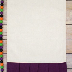 May include: A white kitchen towel with a purple ruffled bottom. The towel is on a wooden surface.