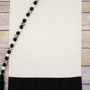 May include: A white kitchen towel with a black ruffled bottom. A black and white beaded garland is draped over the top of the towel.