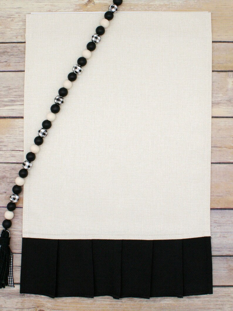 May include: A white kitchen towel with a black ruffled bottom. A black and white beaded garland is draped over the top of the towel.