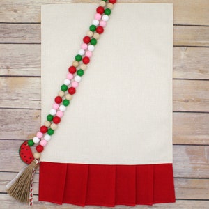 May include: A white kitchen towel with a red ruffled bottom. A watermelon-shaped bead with a brown tassel hangs from a strand of wooden beads in red, pink, green, and natural wood.