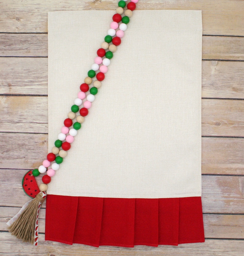 May include: A white kitchen towel with a red ruffled bottom. A watermelon-shaped bead with a brown tassel hangs from a strand of wooden beads in red, pink, green, and natural wood.