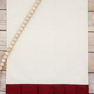 May include: A beige kitchen towel with a red ruffled bottom. The towel is decorated with a strand of wooden beads.