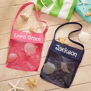May include: Two mesh beach bags, one pink and one navy blue, with the names "Emma Grace" and "Jackson" printed on them in white. The bags are filled with seashells and starfish, and are set on a wooden surface with beach towels.
