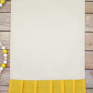 May include: A beige kitchen towel with a yellow ruffled bottom. The towel is on a wood background.