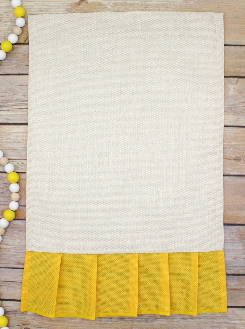 May include: A beige kitchen towel with a yellow ruffled bottom. The towel is on a wood background.