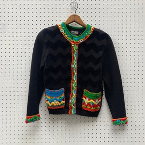Könnte beinhalten: Schwarzer Cardigan mit Chevron-Muster und bunten Perlenbesatz. Der Cardigan hat zwei Taschen mit bunten Perlenbesatz. Der Cardigan hat einen Knopfverschluss und das Label "FANCIES" ist auf der Vorderseite sichtbar.