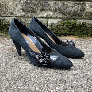 Op de afbeelding: Een paar zwarte suède pumps met een spitse neus en een hoge hak. De schoenen hebben een zwarte roosversiering aan de voorkant.