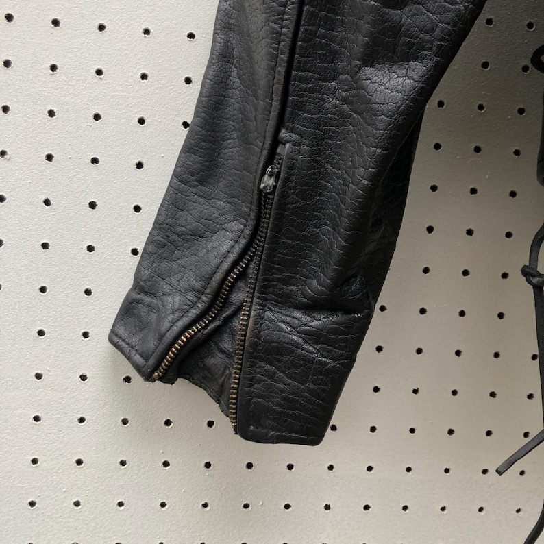 May include: Close-up of a black leather jacket sleeve with a zipper. The zipper is partially open, revealing the lining of the jacket.