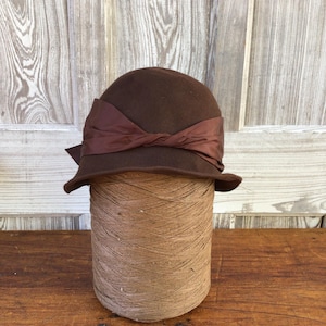 May include: A brown cloche hat with a dark brown ribbon tied in a bow. The hat is displayed on a large spool of tan thread, set against a wooden background. The hat has a rounded crown and a small brim.