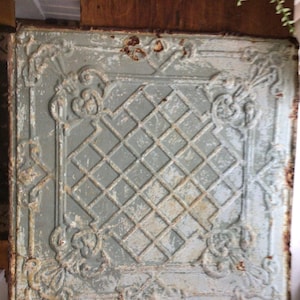 May include: Square, antique-style metal ceiling tile with a distressed, light blue-gray finish. The tile features a diamond grid pattern in the center and ornate floral and scrollwork designs in each corner. The edges show signs of rust, adding to its vintage appeal.