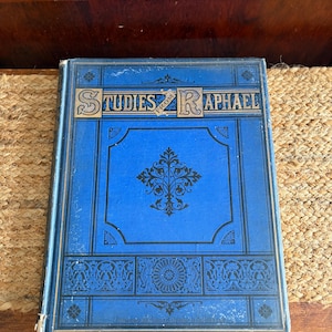 May include: A vintage book titled "Studies from Raphael" with a blue cover and gold lettering. The cover features ornate black and gold designs. The book is resting on a woven surface.
