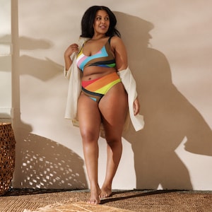 May include: A woman wearing a colourful striped bikini top and matching high-waisted bikini bottoms. She is standing in front of a light beige wall and wearing a white shirt over the bikini.