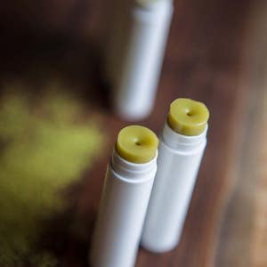 May include: Close-up of two white lip balm tubes with green balm inside. The tubes are on a wooden surface.