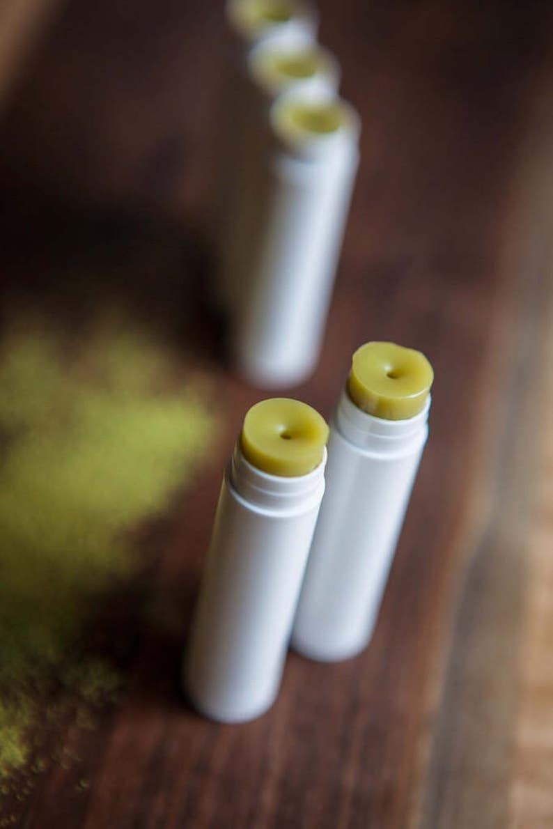 May include: Close-up of two white lip balm tubes with green balm inside. The tubes are on a wooden surface.
