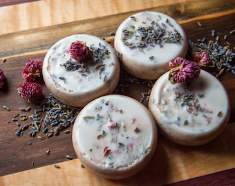 May include: Five round white soap bars with dried lavender and pink flowers on a wooden surface.