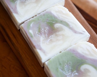 May include: Three rectangular bars of soap featuring a white base with swirling patterns of sage green and soft lavender. Each bar is embossed with a delicate, detailed sprig of lavender on the surface, creating a textured and layered appearance.