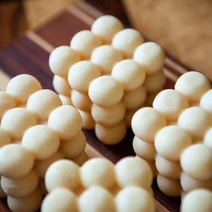 May include: Close-up of several cube-shaped arrangements of small, round, off-white objects. The objects are arranged in a grid-like pattern, creating a textured surface. The background is a wooden cutting board with dark brown and light brown stripes.
