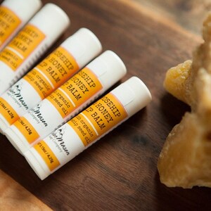 K&ouml;nnte beinhalten: Nahaufnahme von sechs wei&szlig;en Tuben mit Rosehip Honey Lip Balm mit orangenen Etiketten. Die Tuben sind in einer Reihe auf einer Holzoberfl&auml;che angeordnet. Im Hintergrund ist ein Block Bienenwachs zu sehen.