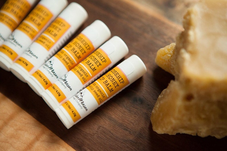 K&ouml;nnte beinhalten: Nahaufnahme von sechs wei&szlig;en Tuben mit Rosehip Honey Lip Balm mit orangenen Etiketten. Die Tuben sind in einer Reihe auf einer Holzoberfl&auml;che angeordnet. Im Hintergrund ist ein Block Bienenwachs zu sehen.
