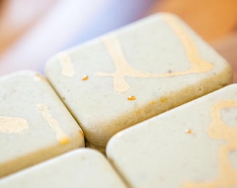 May include: Four square, light sage green tiles with rounded corners, each featuring a unique, hand-painted metallic gold abstract design on the surface. The tiles have a smooth, matte finish and are arranged in a tight, symmetrical grid pattern.