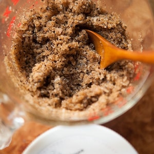 May include: A close-up of a glass measuring cup filled with a brown coffee scrub. The scrub is made with coffee grounds and sugar. A wooden spoon is partially submerged in the scrub.