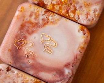 May include: Three rectangular bars of soap with a marbled pink and white surface. Each bar features embedded coarse amber-colored salt crystals and small, gold-colored embossed heart outlines stamped into the smooth soap surface.