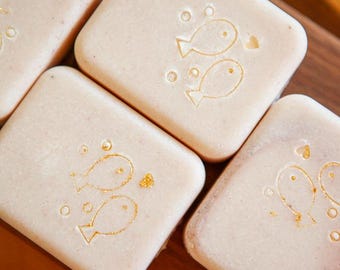 May include: Several rectangular bars of light beige soap with rounded corners. Each bar features a stamped design of two small fish, bubbles, and a heart, with some of the stamped details highlighted in a metallic gold color.