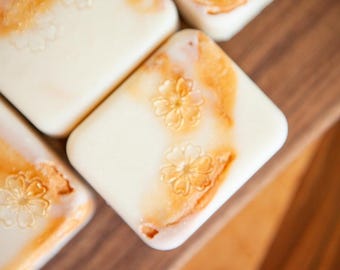 May include: Rectangular off-white soap bars featuring a smooth surface with embossed flower patterns. Each bar displays a marbled orange and peach-colored swirl design across the top, creating a distinct contrast against the creamy white base.