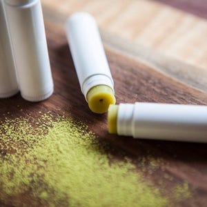 May include: A close-up of four white lip balm tubes with a yellow lip balm inside. The lip balms are on a wooden surface with green powder scattered around them.