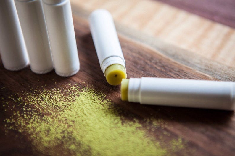 May include: A close-up of four white lip balm tubes with a yellow lip balm inside. The lip balms are on a wooden surface with green powder scattered around them.