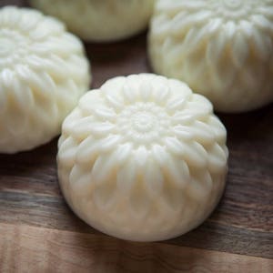 Puede incluir: Cuatro jabones decorativos redondos y blancos con un diseño floral. Los jabones están dispuestos sobre una superficie de madera.