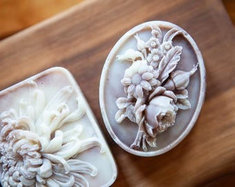 May include: Two decorative soaps with floral designs. One is oval-shaped with a bouquet of flowers, including roses and daisies. The other is rectangular with a chrysanthemum. Both soaps are light purple and white.