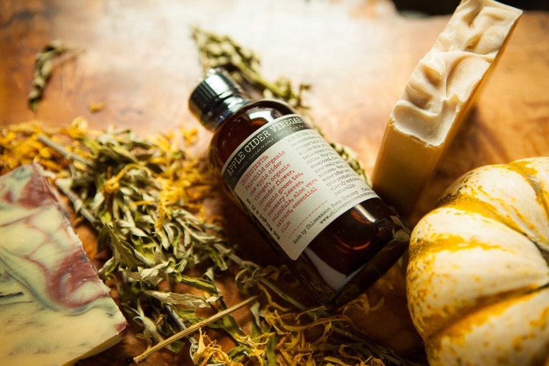 May include: A brown glass bottle with a label that reads "Apple Cider Vinegar" and lists ingredients including "fermented raw apple cider, organic ginger, organic rosemary flower, organic green tea, organic green leaves, calendula, aloe vera, citrus essential oil". The bottle is sitting on a wooden surface with dried flowers and a small white pumpkin.