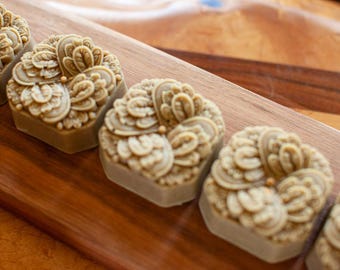 May include: A series of light brown, hexagonal-shaped soaps featuring an intricate, raised floral pattern on the top surface. Each soap has a smooth, flat base and a detailed, textured design consisting of curved, petal-like shapes and small dots.
