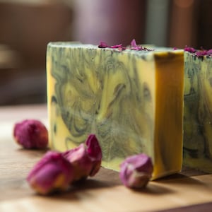 May include: Two rectangular bars of handmade soap with a yellow base and swirling black patterns. Dried rose petals are scattered on top of the soap and around the base. The soap is on a wooden surface.