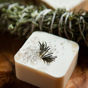 Puede incluir: Una barra de jabón blanca con una ramita de romero y purpurina plateada en la parte superior. El jabón está sobre una superficie de madera.