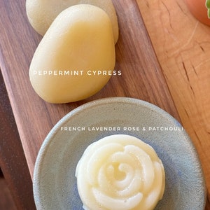 Puede incluir: Dos barras de jabón redondas de color amarillo pálido sobre una superficie de madera. La barra de jabón de la izquierda está etiquetada como "Peppermint Cypress". Una pequeña barra de jabón redonda de color amarillo pálido con forma de rosa está sobre un plato de cerámica azul y gris. La barra de jabón está etiquetada como "French Lavender Rose & Patchouli".