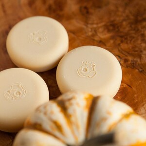 May include: Three round, white soap bars with a small animal design on each bar. The soap bars are sitting on a wooden surface next to a white pumpkin.