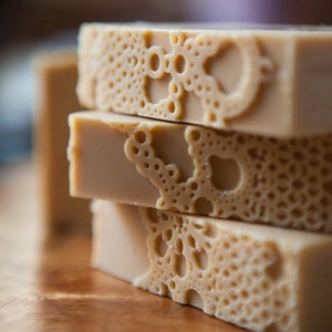Pu&ograve; includere: Una pila di quattro saponette fatte a mano con un motivo decorativo di cerchi e volute. Le saponette sono di colore beige chiaro.