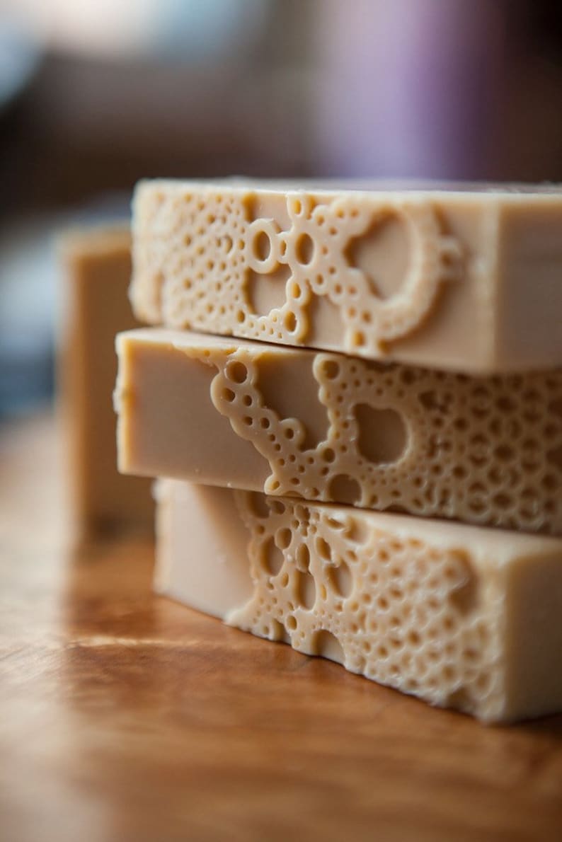 Pu&ograve; includere: Una pila di quattro saponette fatte a mano con un motivo decorativo di cerchi e volute. Le saponette sono di colore beige chiaro.