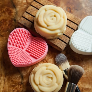 May include: Two round, white, rose-shaped soaps on a wooden soap dish. A pink heart-shaped silicone facial cleansing brush and a makeup brush are also pictured.