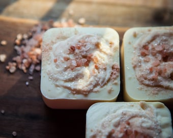 Pode incluir: Close-up de três sabonetes quadrados com base branca cremosa, cobertos com um redemoinho de creme texturizado e salpicados com cristais de sal rosa do Himalaia. Os sabonetes estão dispostos sobre uma superfície de madeira escura.