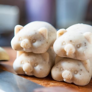 May include: Four white, pig-shaped soaps stacked on a wooden surface. The soaps have a textured surface and are slightly off-white in color.