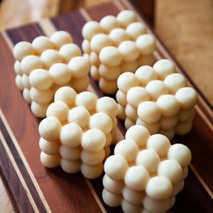 May include: Five ivory-colored, cube-shaped candles with a bubble design are arranged on a wooden cutting board. The candles are made of small, round spheres clustered together. The cutting board has a striped pattern of dark and light brown wood.