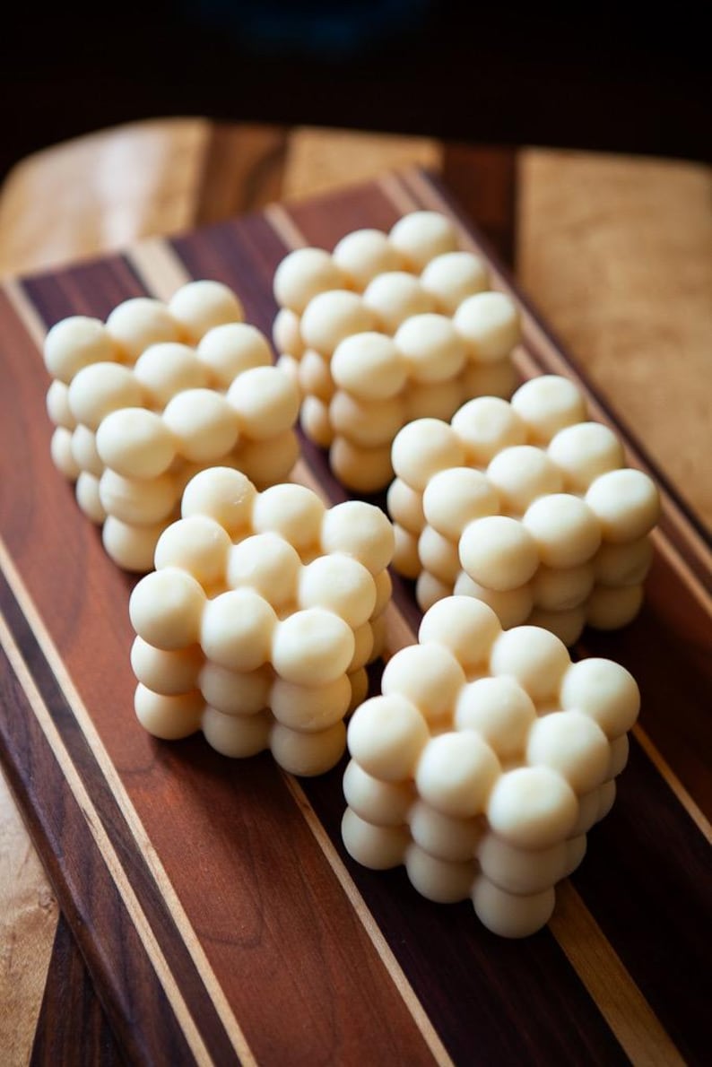 May include: Five ivory-colored, cube-shaped candles with a bubble design are arranged on a wooden cutting board. The candles are made of small, round spheres clustered together. The cutting board has a striped pattern of dark and light brown wood.